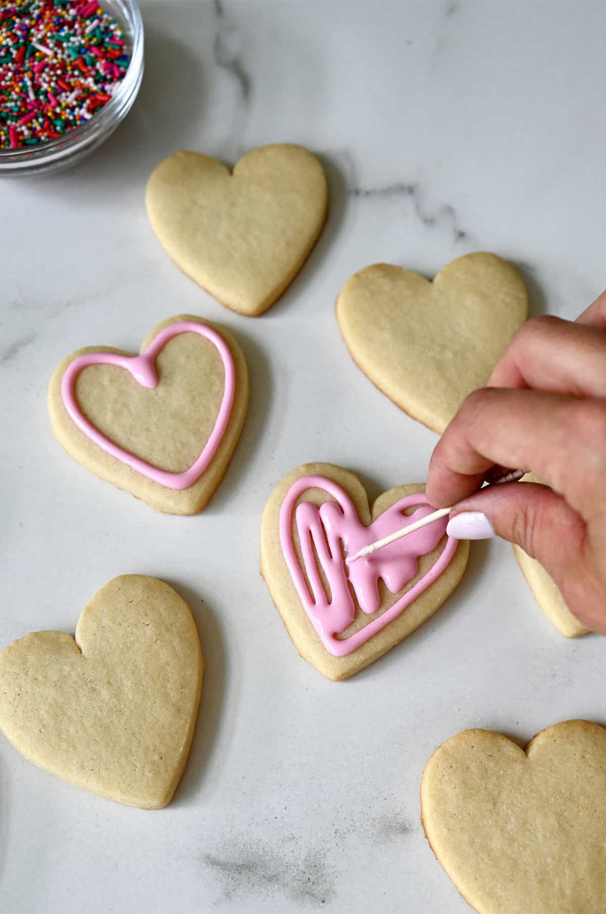 Using a toothpick to smooth out royal icing on a heart-shaped sugar cookie.