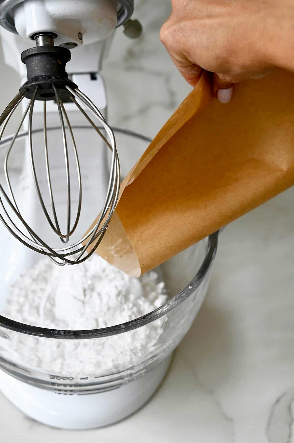 Transferring sifted powdered sugar from parchment paper into a bowl of a stand mixer fitted with the whisk attachment.