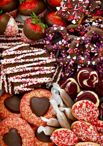Festive Valentine's Day desserts, including Valentine's Day peanut butter blossoms, chocolate sugar cookies, red velvet cheesecake brownie bites, soft caramel candies, chocolate-covered pretzels, and white chocolate dipped strawberries.