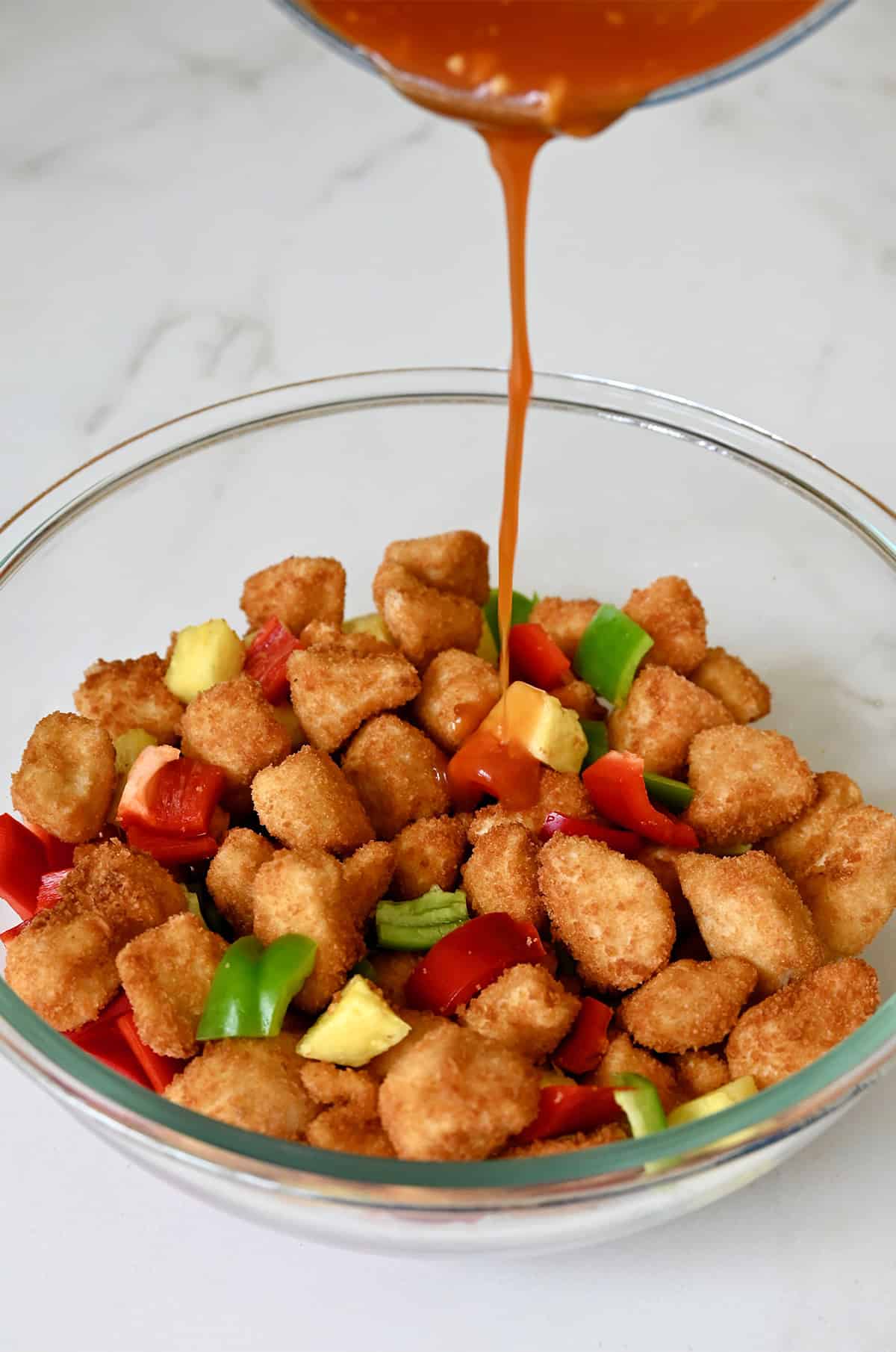 Adding sweet and sour sauce atop fried chicken, pineapple chunks, and red and green bell peppers in a bowl.