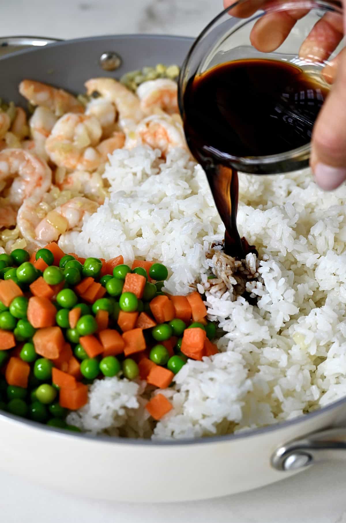 Adding soy sauce to a large skillet with cooked jasmine rice, veggies and shrimp to make fried rice.