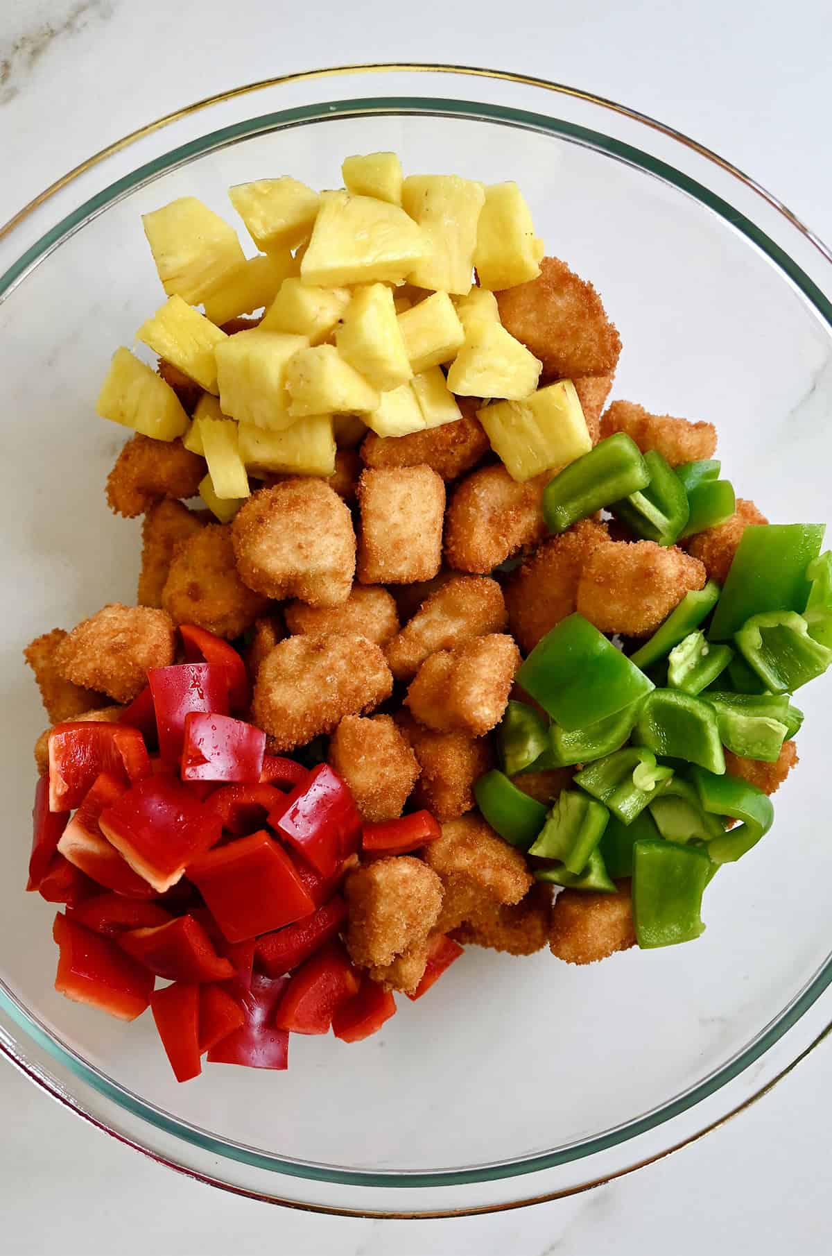A bowl containing battered fried chicken, pineapple chunks, and chopped red and green bell peppers.