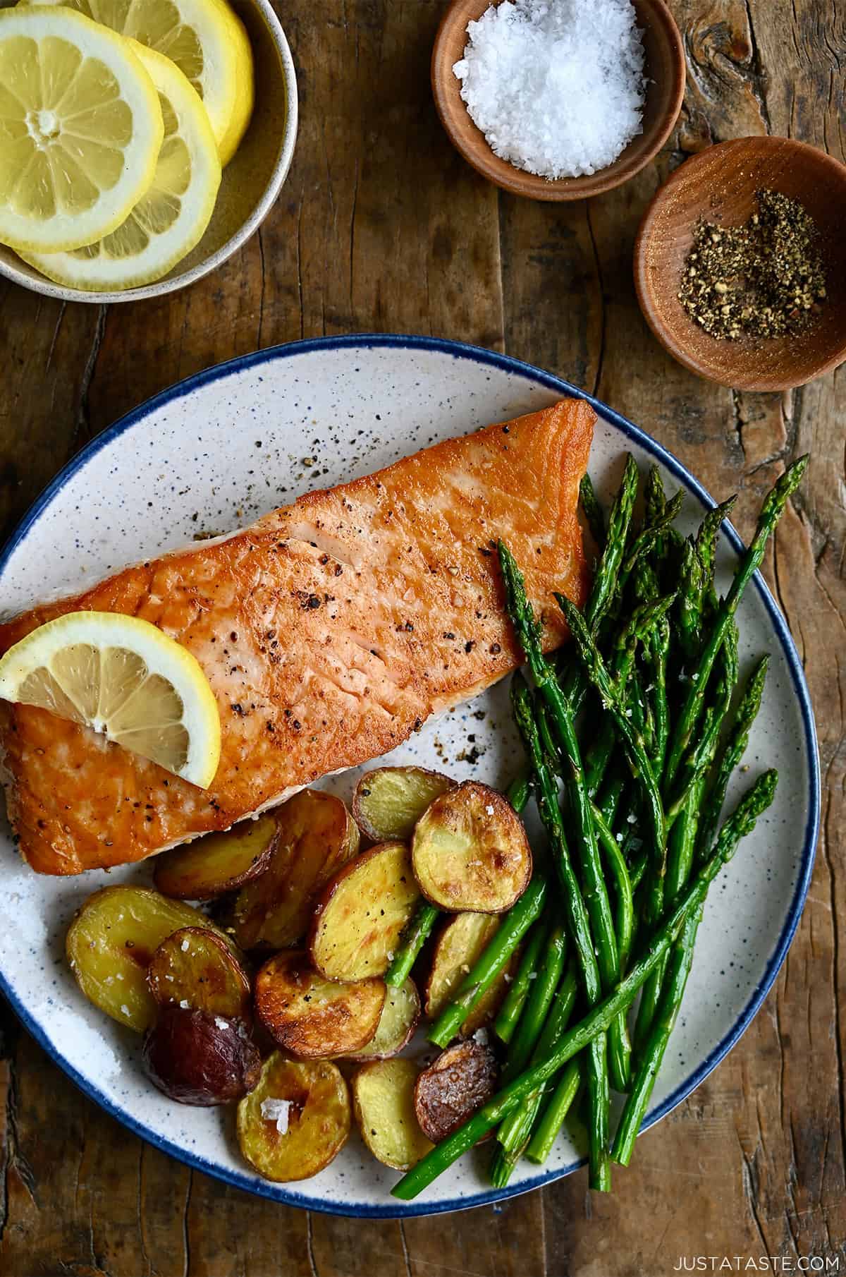 A pan seared salmon filet with a golden crust, topped with a lemon slice on a plate with roasted baby potatoes and asparagus spears.