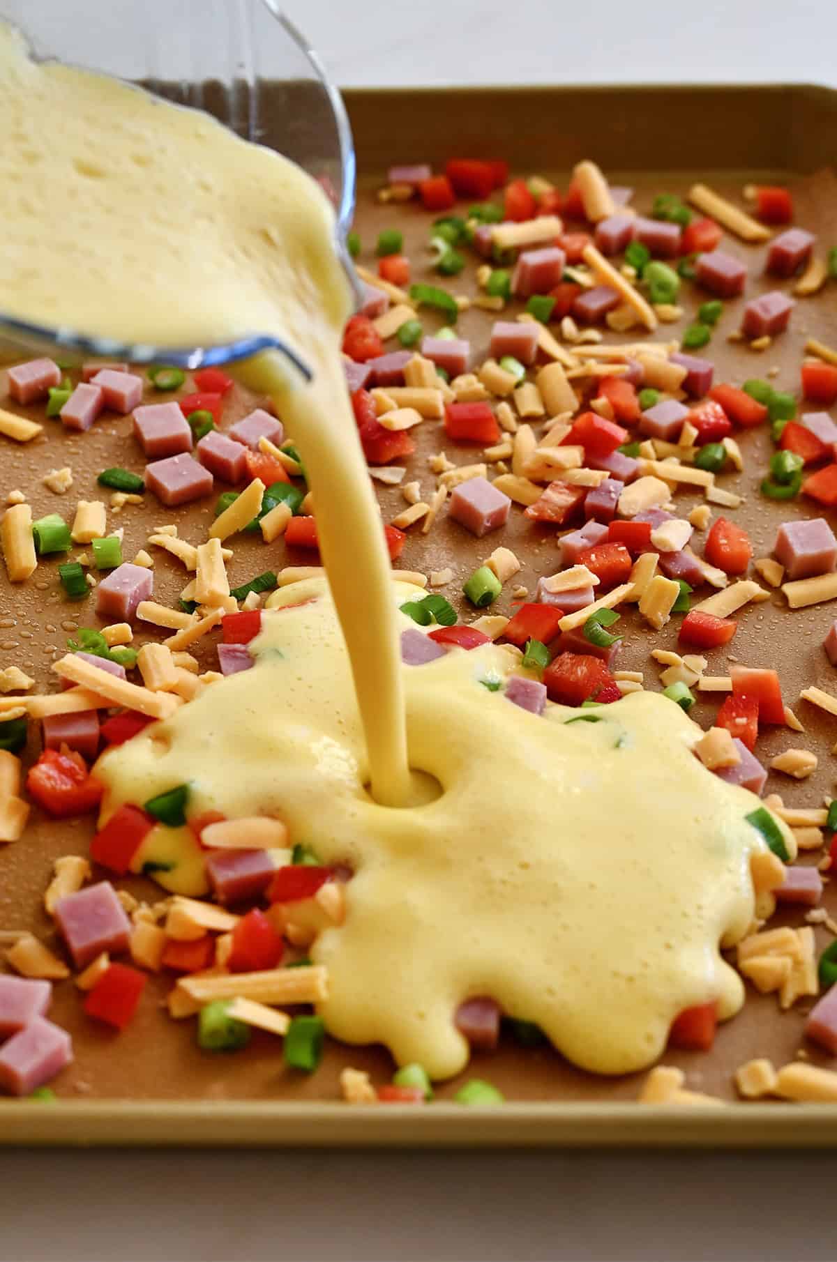 Pouring whipped eggs from a blender onto a sheet pan with diced veggies, ham, and shredded cheese.