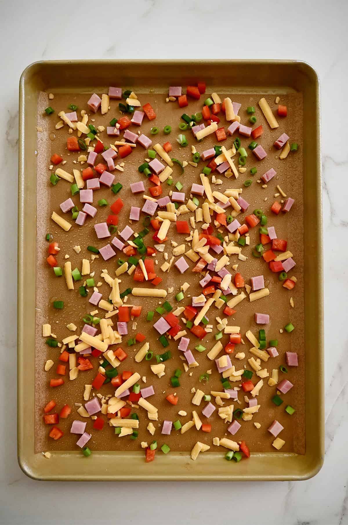 Diced bell peppers, shredded cheddar cheese and diced ham on a parchment paper-lined baking sheet.