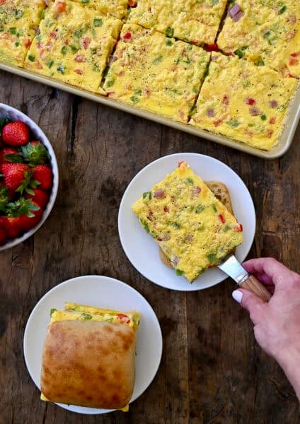 Sheet pan eggs with diced bell peppers, shredded cheddar cheese and diced ham being dished out onto plates with ciabatta rolls to make breakfast sandwiches.