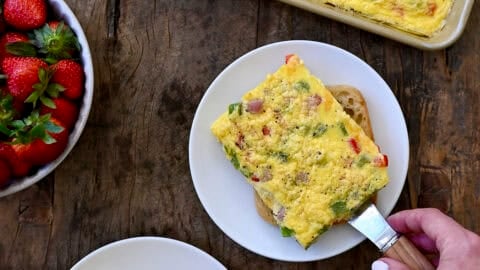 Sheet pan eggs with diced bell peppers, shredded cheddar cheese and diced ham being dished out onto plates with ciabatta rolls to make breakfast sandwiches.