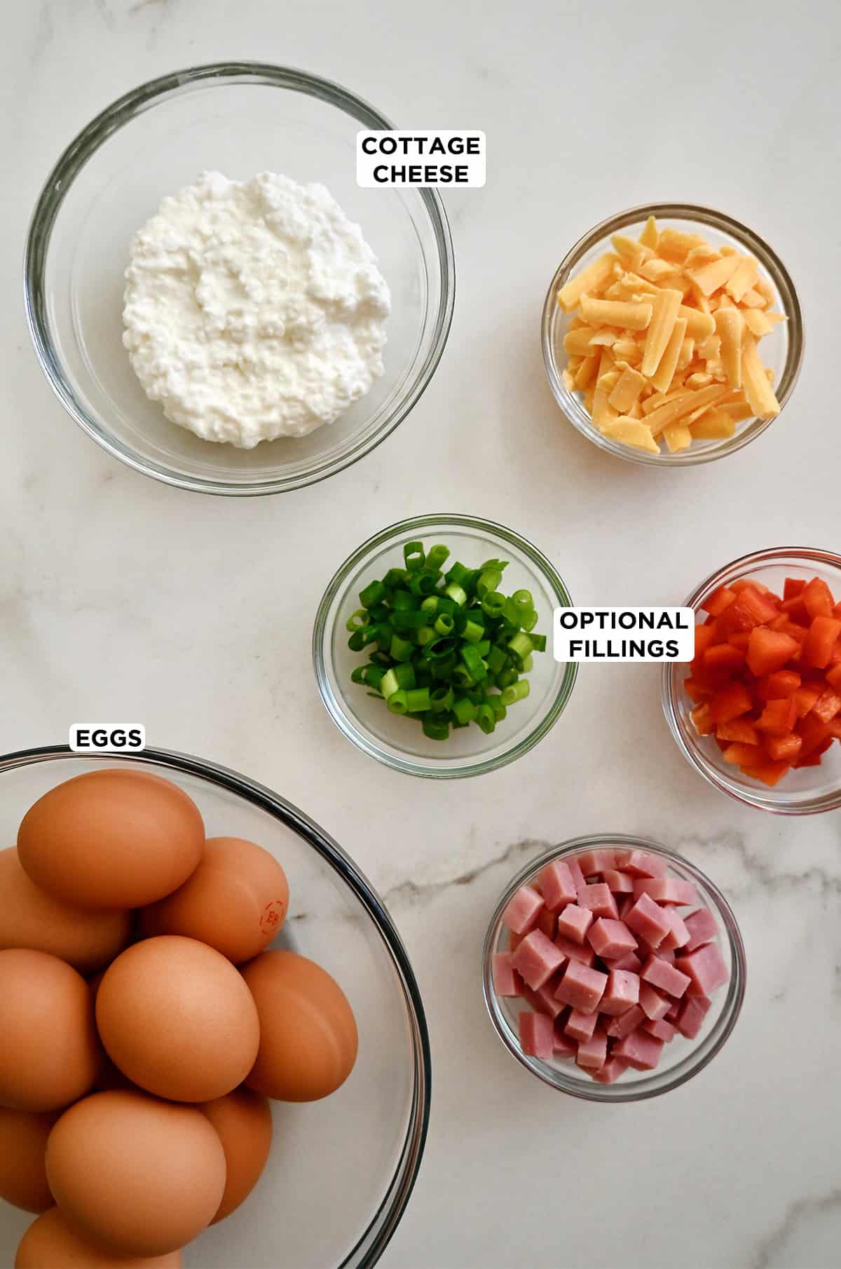 Ingredients to make sheet pan eggs with cottage cheese and optional add-ins, including diced bell peppers, shredded cheddar cheese and diced ham.