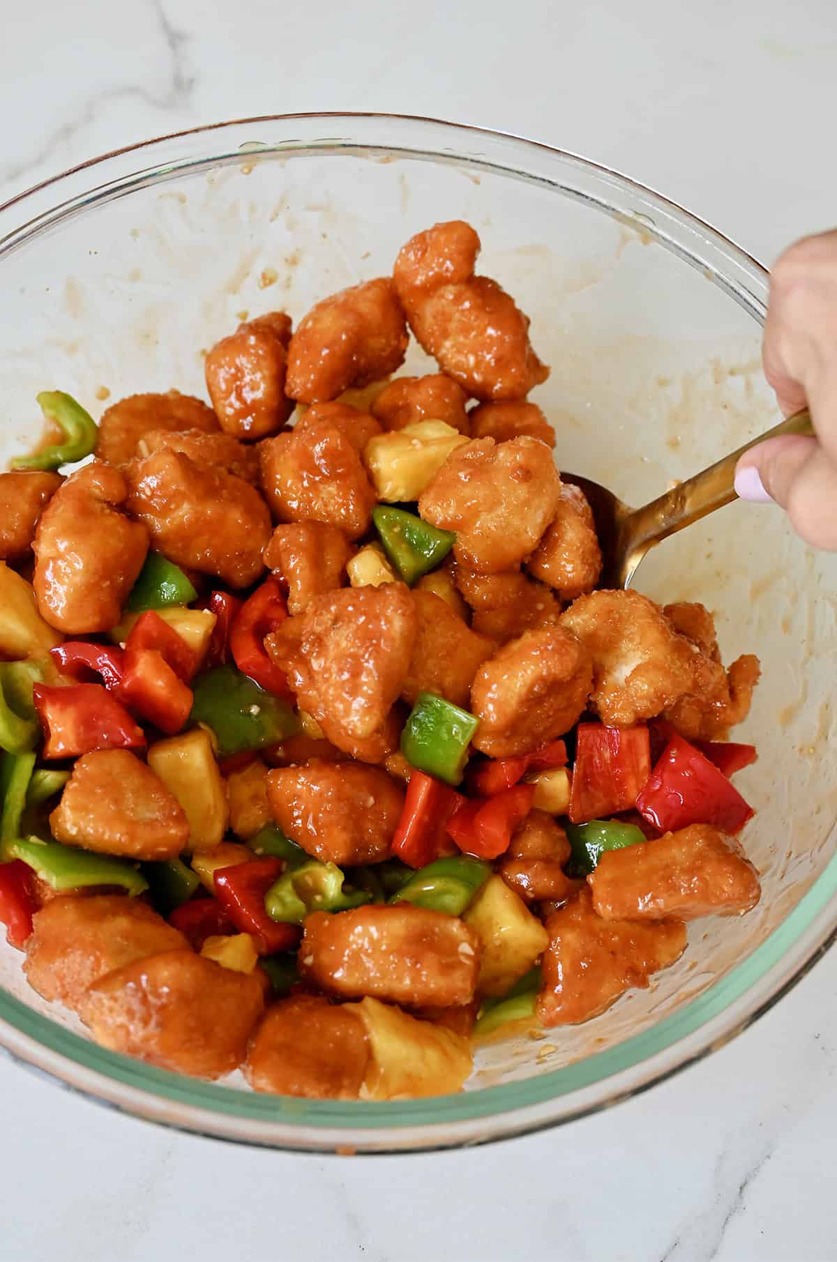 Tossing fried chicken, pineapple, and bell peppers in a homemade sweet and sour sauce in a bowl.