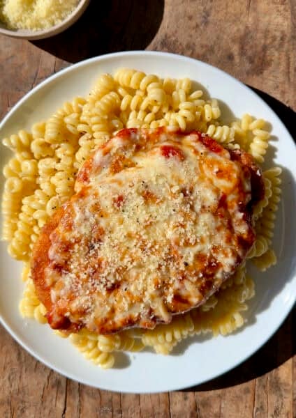 Chicken parmesan (a crispy breaded chicken cutlet topped with marinara sauce and melted mozzarella cheese) atop cacio e pepe pasta on a dinner plate.