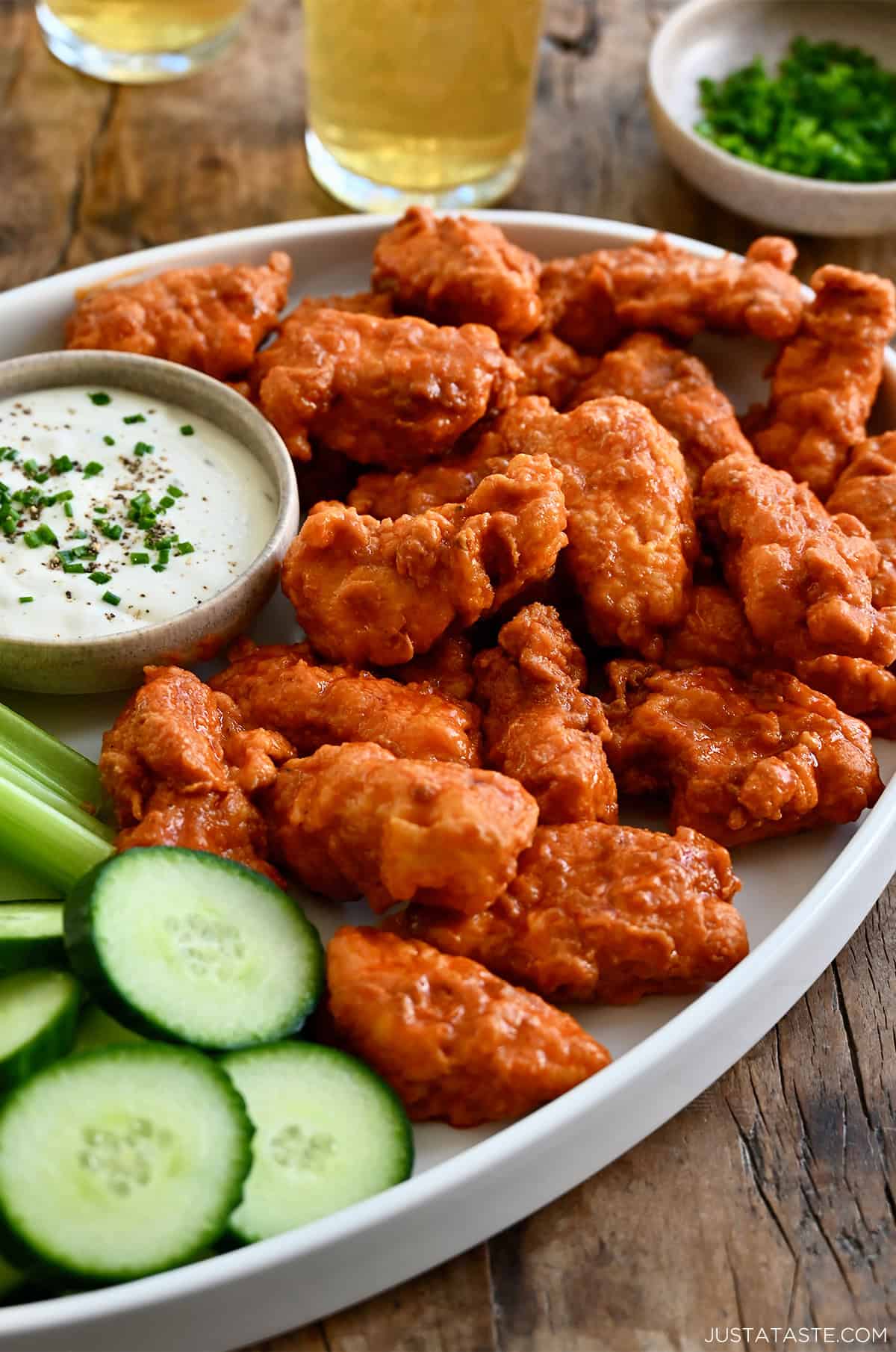 Boneless chicken wings tossed in Buffalo sauce on a serving platter with a small bowl of blue cheese dressing, celery sticks and cucumber slices.