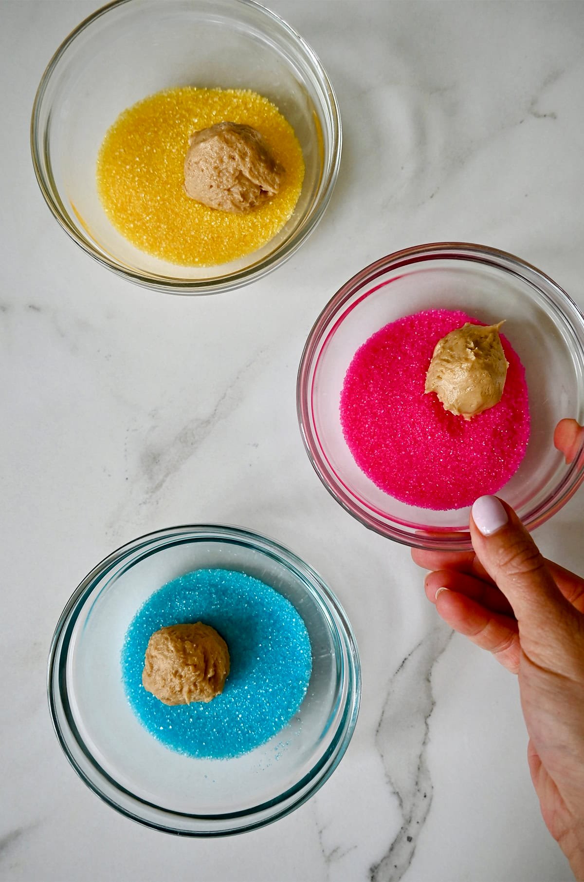 Rolling round balls of peanut butter cookie dough in small bowls containing pink, blue and yellow sanding sugar.