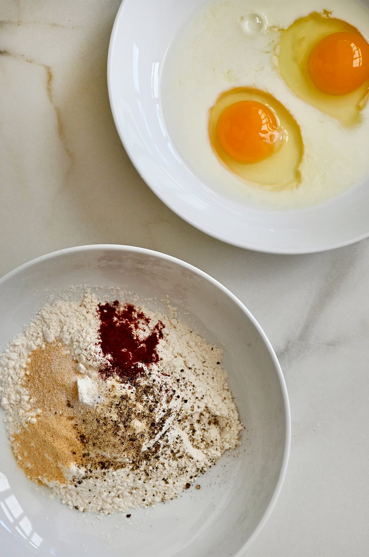 Two shallow bowls, one containing flour and seasonings, the other containing two eggs.