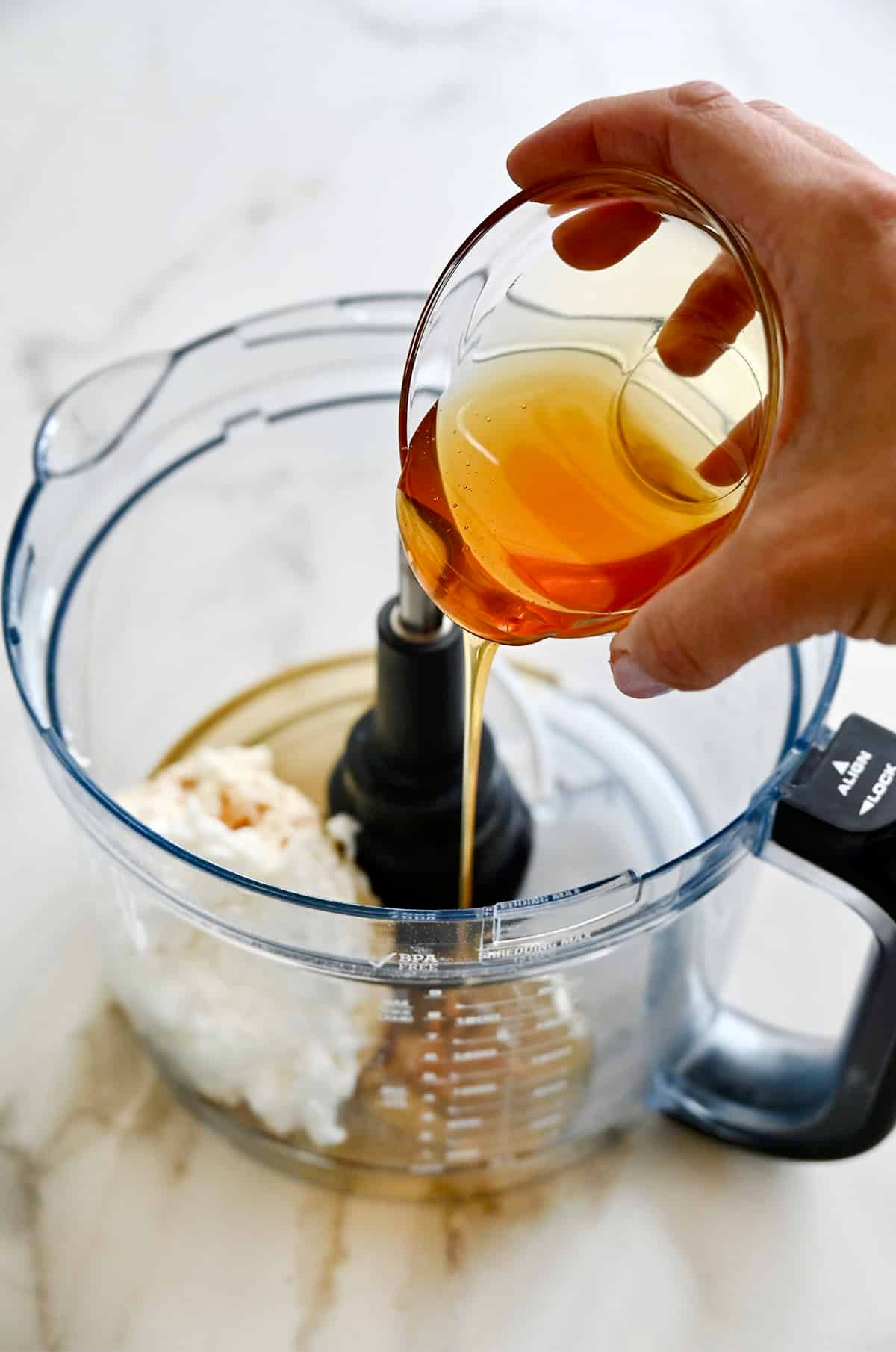 Pouring honey into a food processor bowl with cottage cheese and vanilla extract.