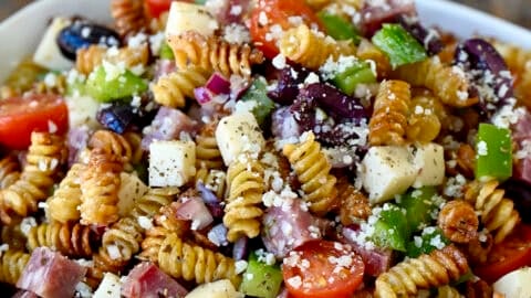 Crispy pasta salad with cubed provolone, salami, olives, tomatoes and diced bell peppers in a large bowl.