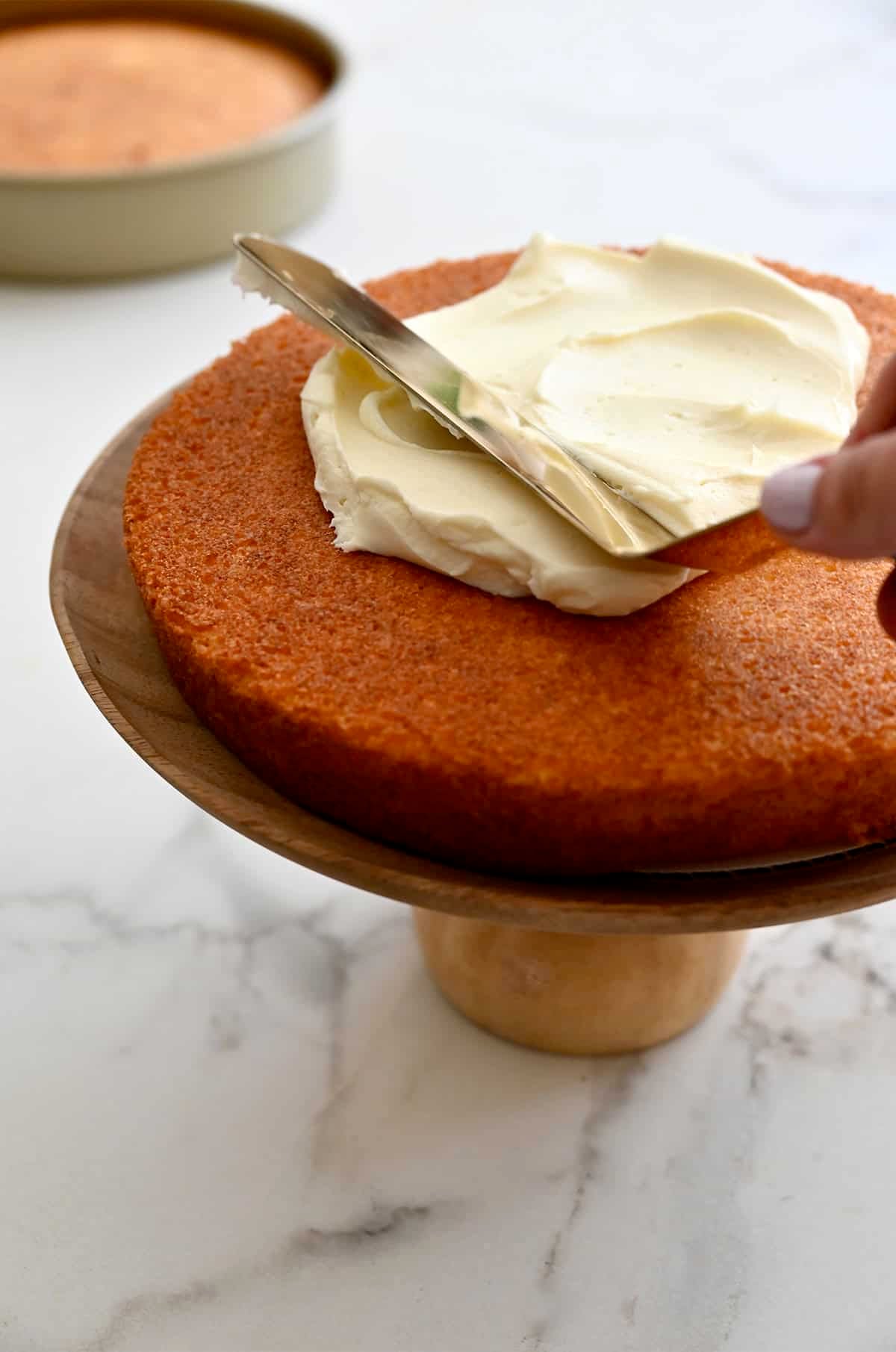 Using an offset spatula to spread cream cheese frosting onto the first layer of a two-tiered coconut cake.