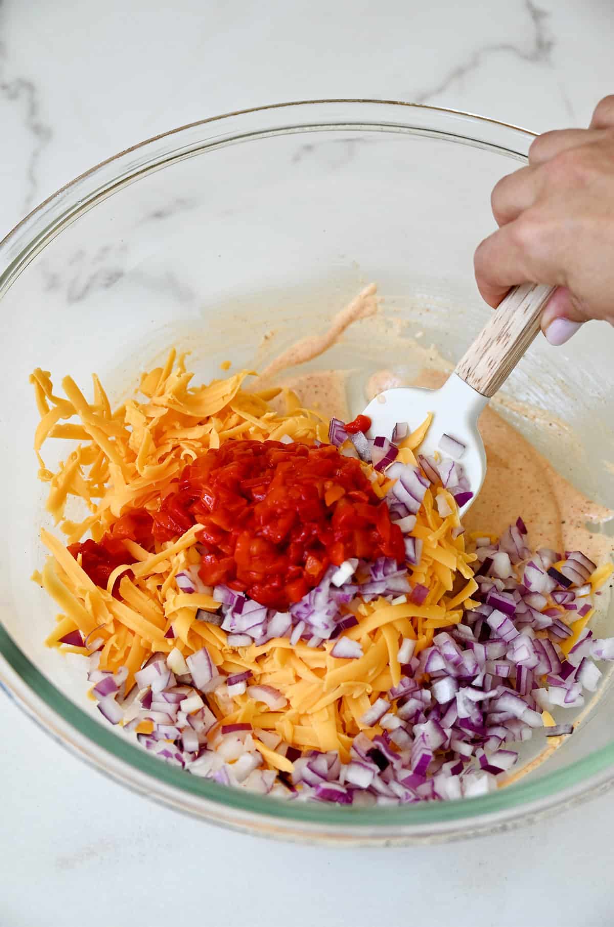 Mixing shredded cheddar, pimento peppers, diced red onions, mayonnaise and spices in a large glass bowl to make a dressing for pasta salad.