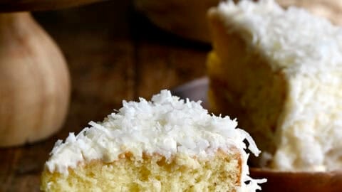 A slice of moist coconut cake with plenty of cream cheese frosting and flaked coconut on a plate.