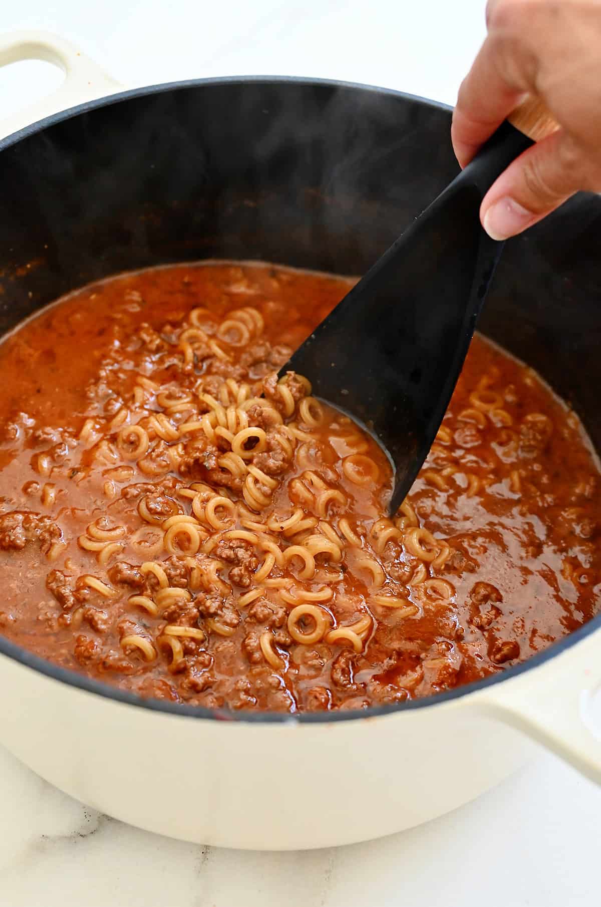 Adding anelli pasta to a Dutch oven with tomato meat sauce.