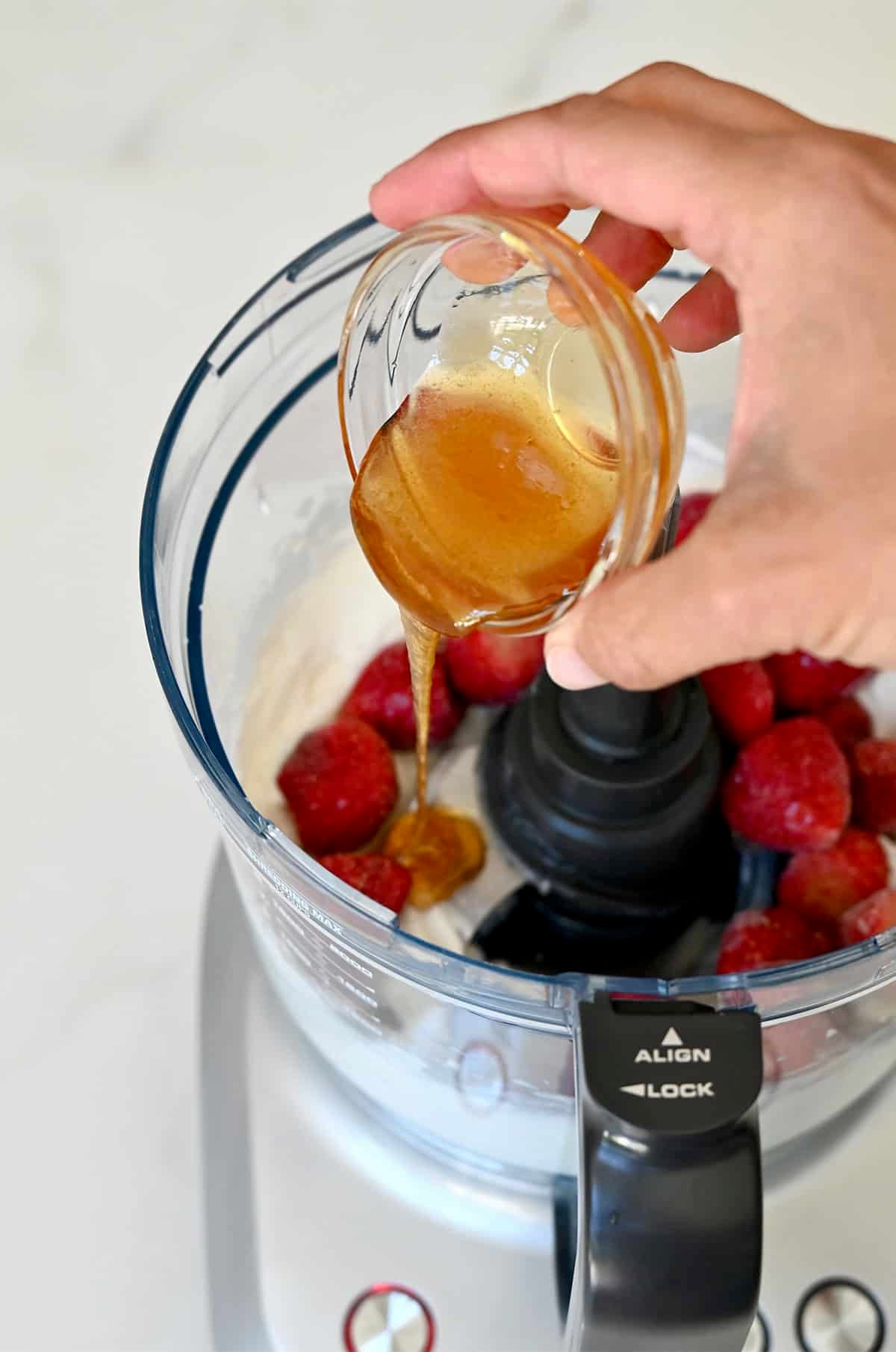 Adding honey from a small bowl into a food processor with blended cottage cheese and frozen fruit.