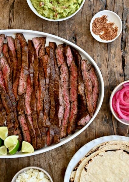 Sliced carne asada on a serving plate surrounded by plates containing tortillas, guacamole, pickled onions and cotija cheese.