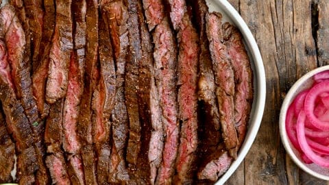 Sliced carne asada on a serving plate surrounded by plates containing tortillas, guacamole, pickled onions and cotija cheese.
