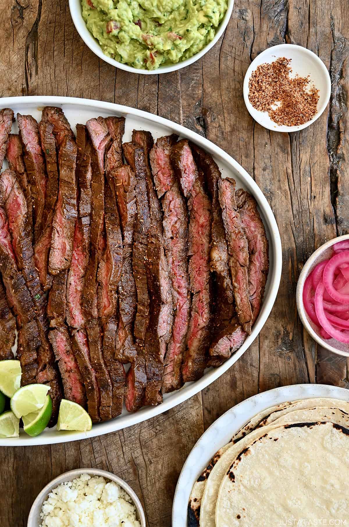Sliced carne asada on a serving plate surrounded by plates containing tortillas, guacamole, pickled onions and cojita cheese.