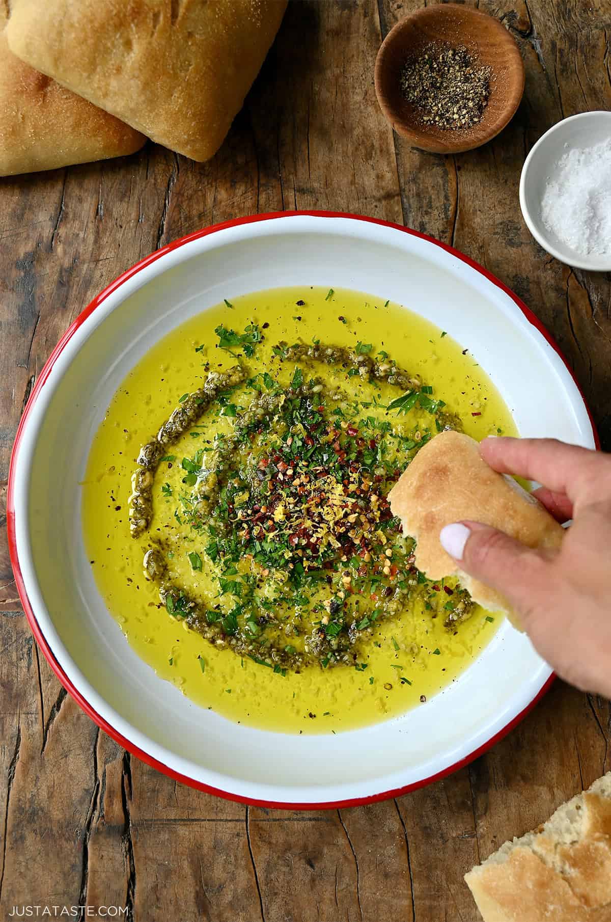 A hand holding a chunk of bread dipping into olive oil with pesto and herbs on a shallow plate.