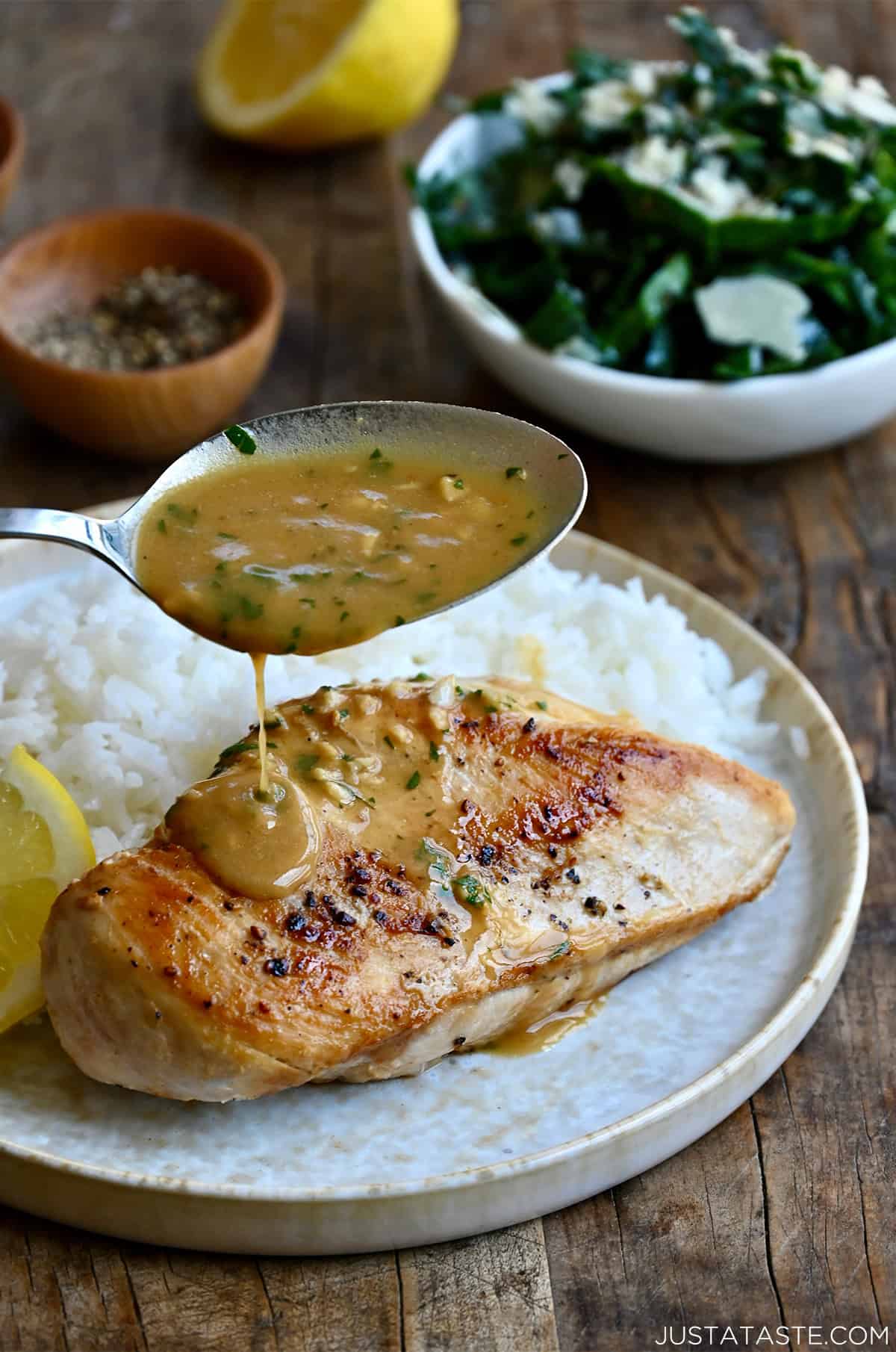 Spooning garlic butter pan sauce atop a seared chicken breast on a plate with white rice and a lemon slice.