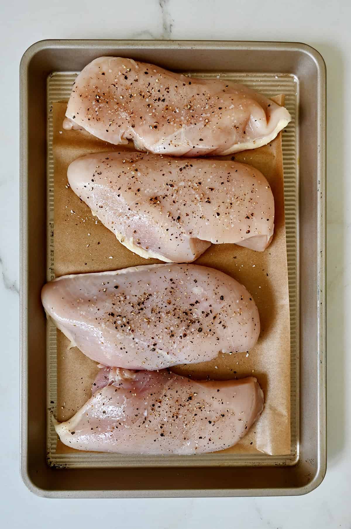 Salt and pepper seasoned chicken breasts on a parchment paper-lined baking sheet before pan-frying.