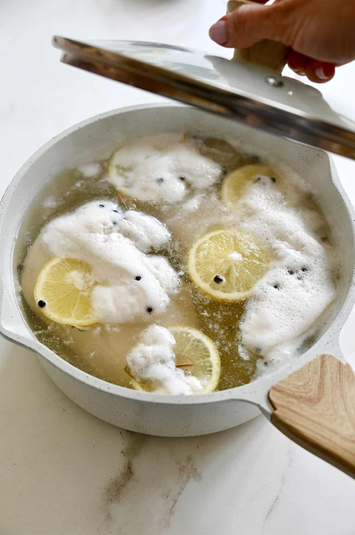 Poached chicken in a pot with lemon slices, peppercorns, garlic cloves and bay leaves.
