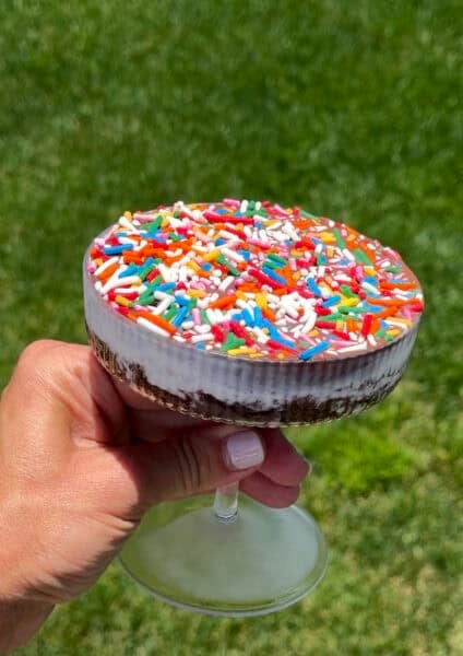 A hand holds an ice cream cake cocktail featuring a layer of chocolate cake topped with vanilla ice cream, Magic Shell and rainbow sprinkles.