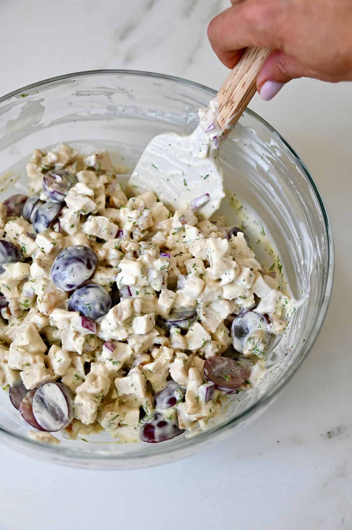 Mixing chicken salad with grapes and walnuts in a creamy mayo-based dressing in a large bowl.