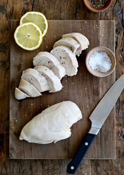 Poached chicken topped with flaky sea salt and cracked black pepper on a wooden cutting board with lemon slices, a small bowl containing salt and a chef's knife.