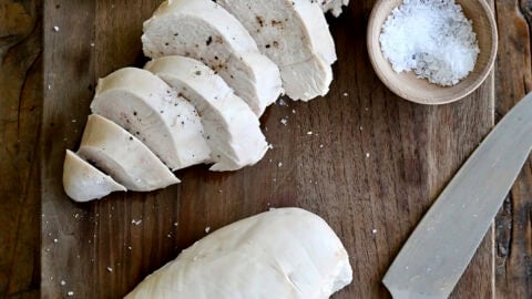 Poached chicken topped with flaky sea salt and cracked black pepper on a wooden cutting board with lemon slices, a small bowl containing salt and a chef's knife.