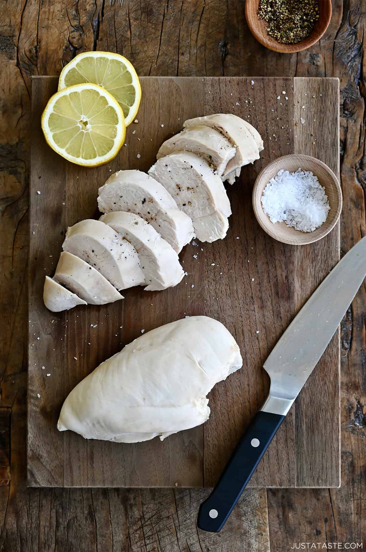 Poached chicken topped with flaky sea salt and cracked black pepper on a wooden cutting board with lemon slices, a small bowl containing salt and a chef's knife.