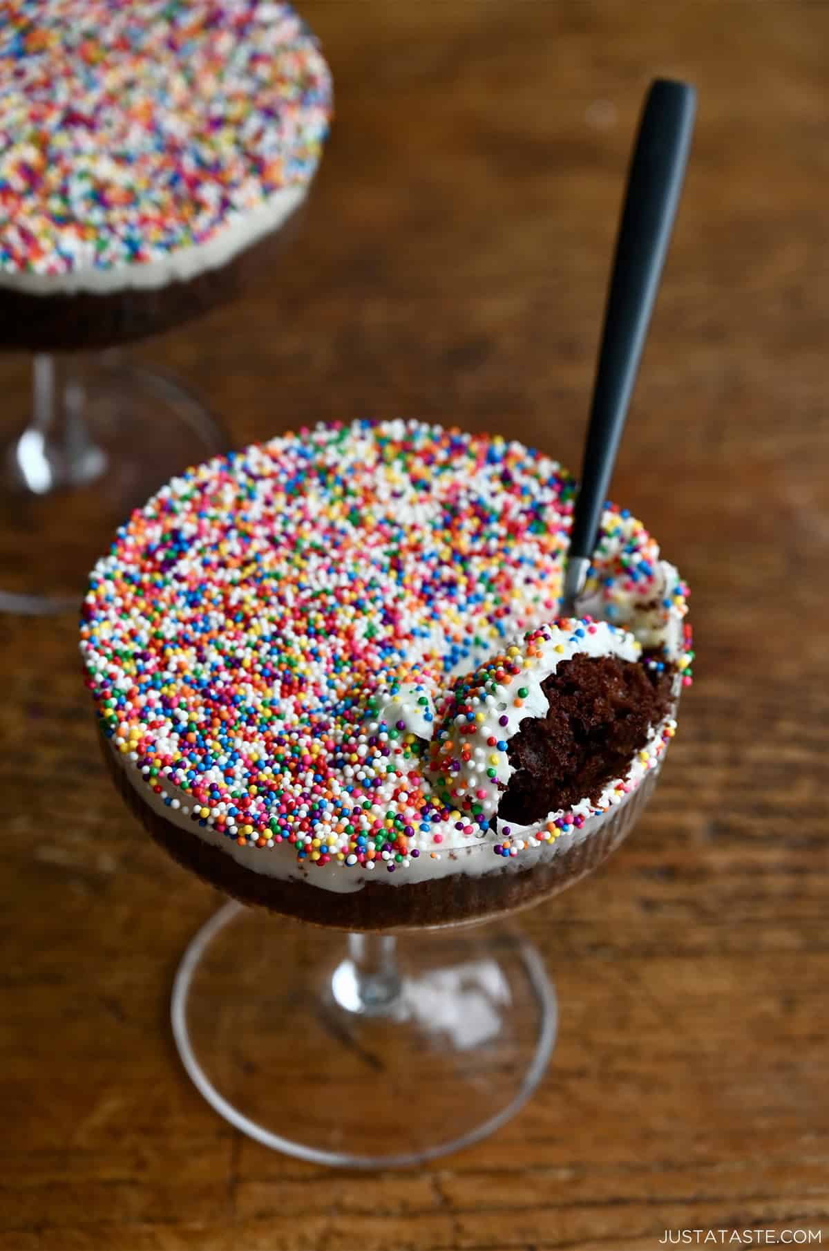 A spoon scooping out cake and frosting with rainbow nonpareils from a cocktail glass.