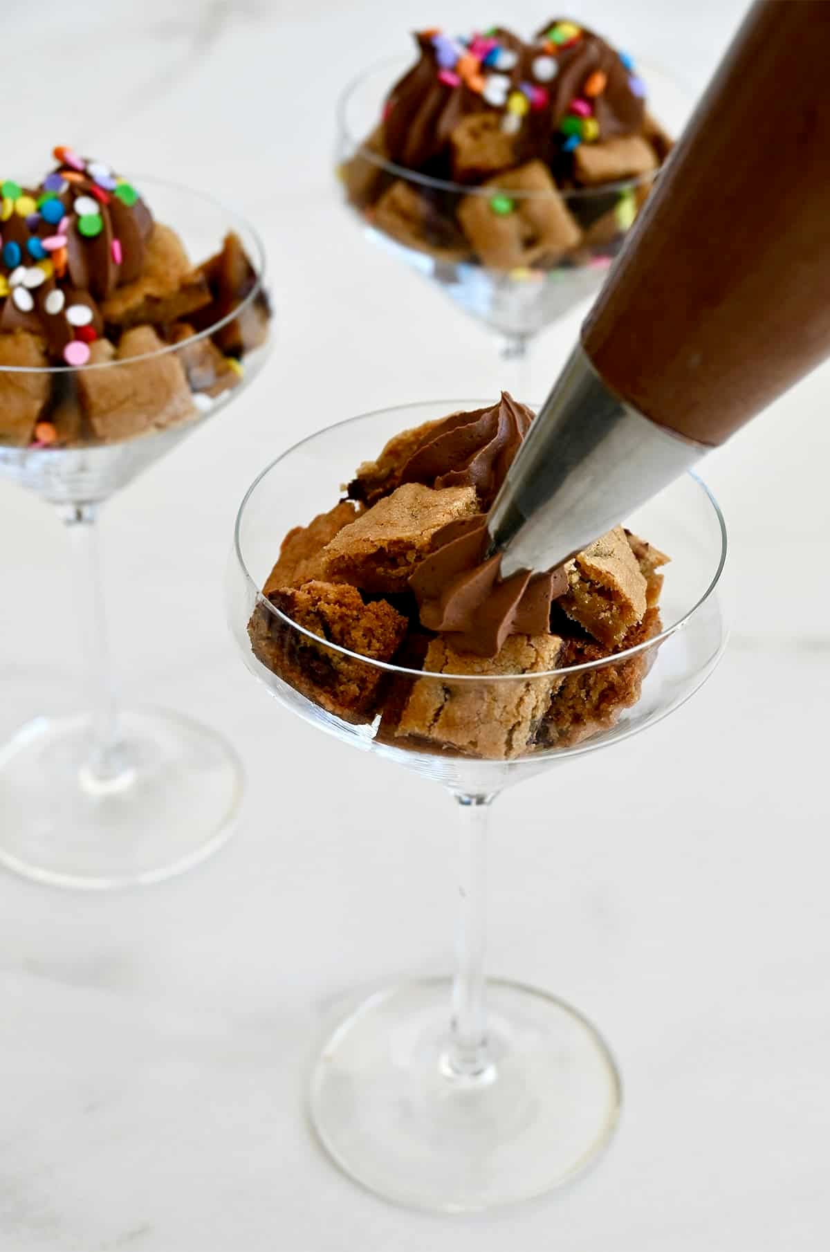 Piping chocolate buttercream frosting atop cookie cubes in a cocktail glass.