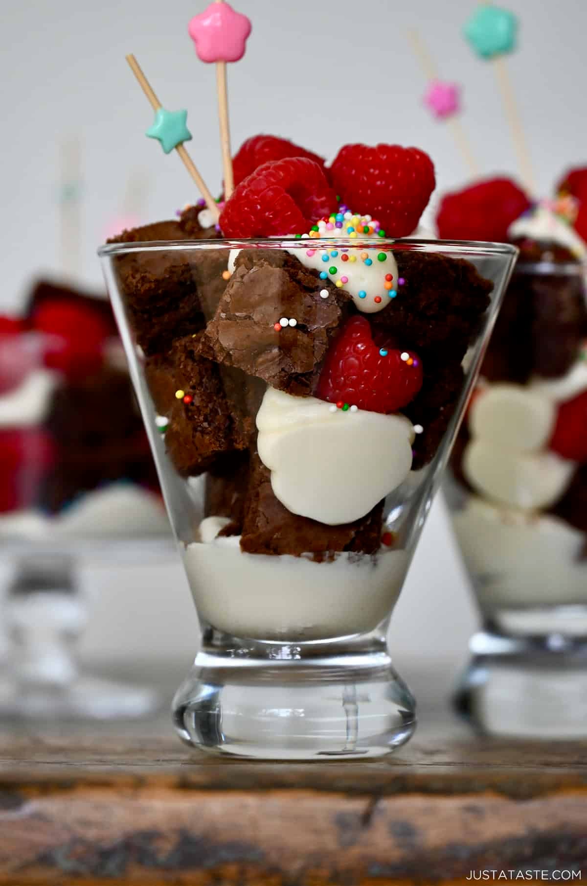 A cocktail glass with layers of buttercream frosting, brownie cubes, raspberries and sprinkles.