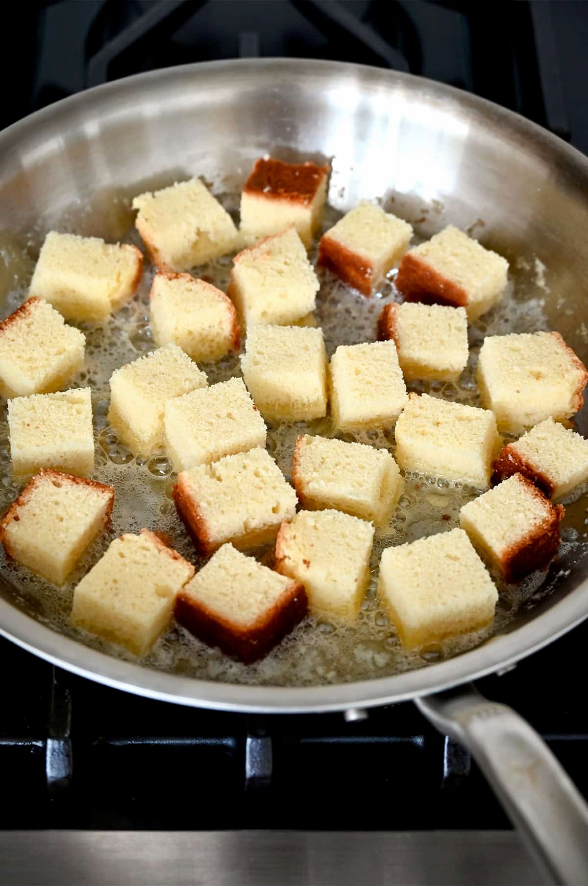 Cubes of pound cake are frying in melted butter in a stainless steel skillet.