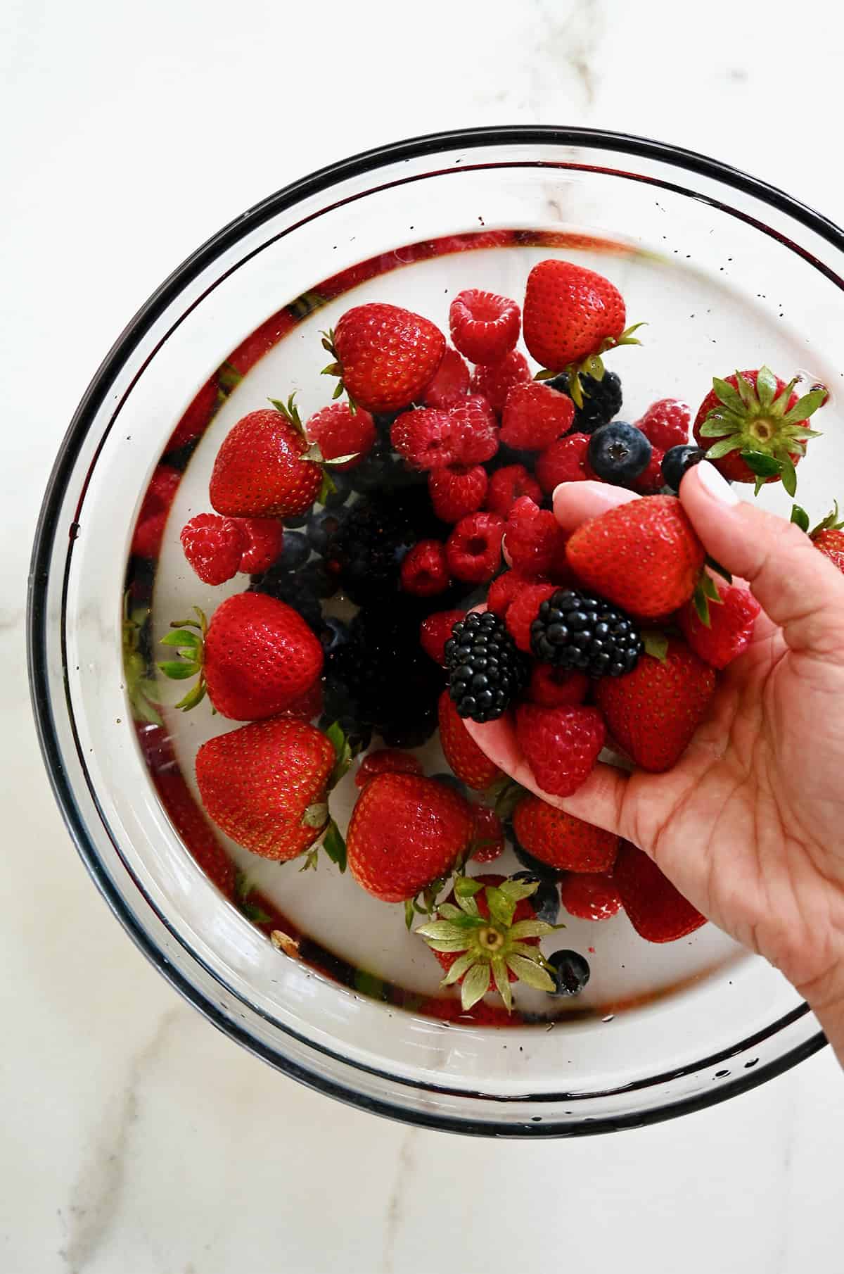 A hand holds fresh berries over a large bowl containing water.