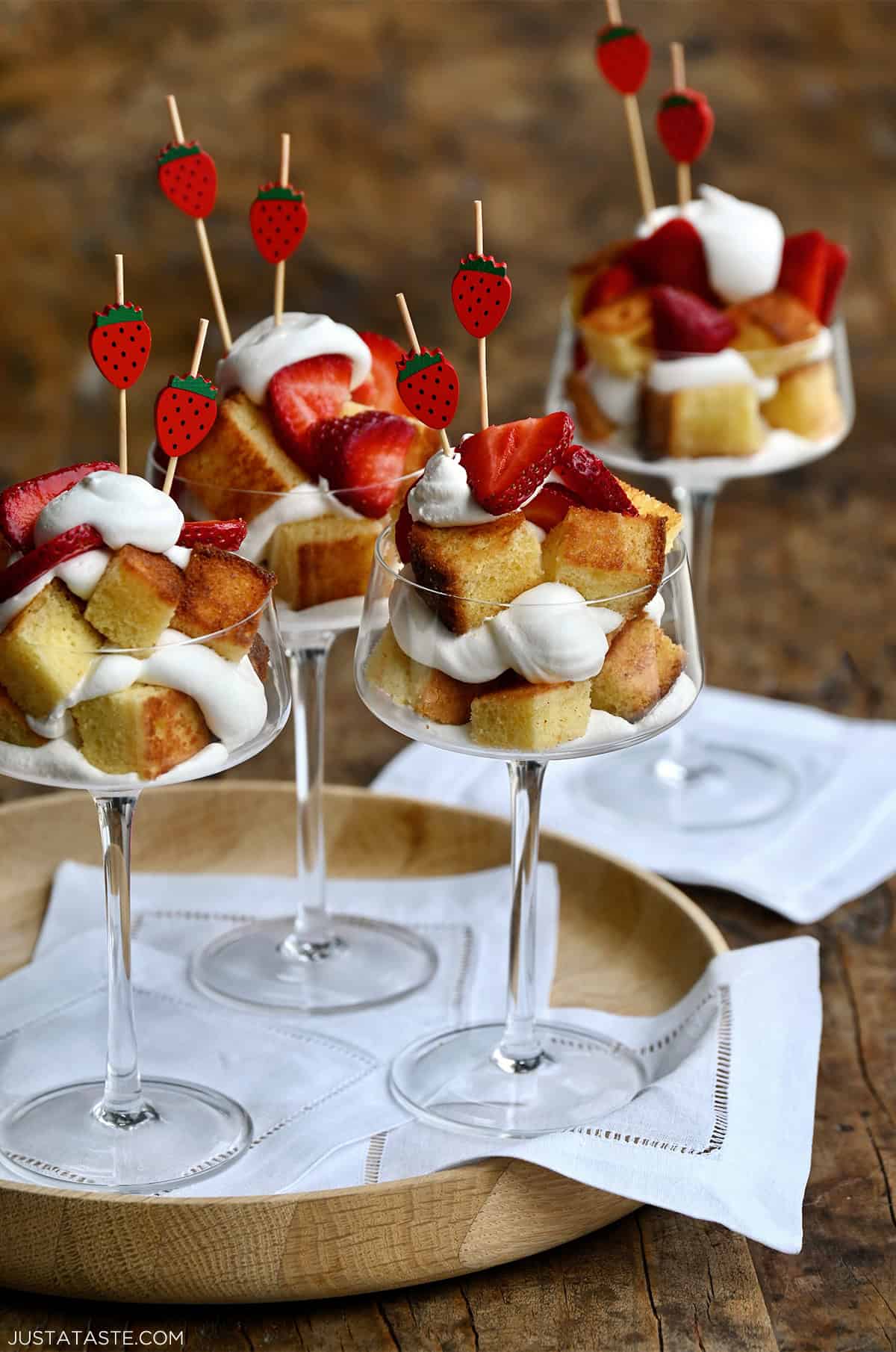 Glass cups filled with layers of toasted pound cake cubes, whipped cream and sliced strawberries, topped with strawberry-shaped toothpicks, served on a wooden tray.