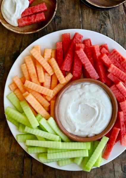 Greek yogurt fruit dip in a small bowl on a plate surrounded by a rainbow of fruit.