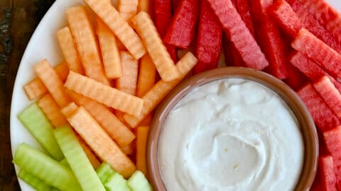 Greek yogurt fruit dip in a small bowl on a plate surrounded by a rainbow of fruit.