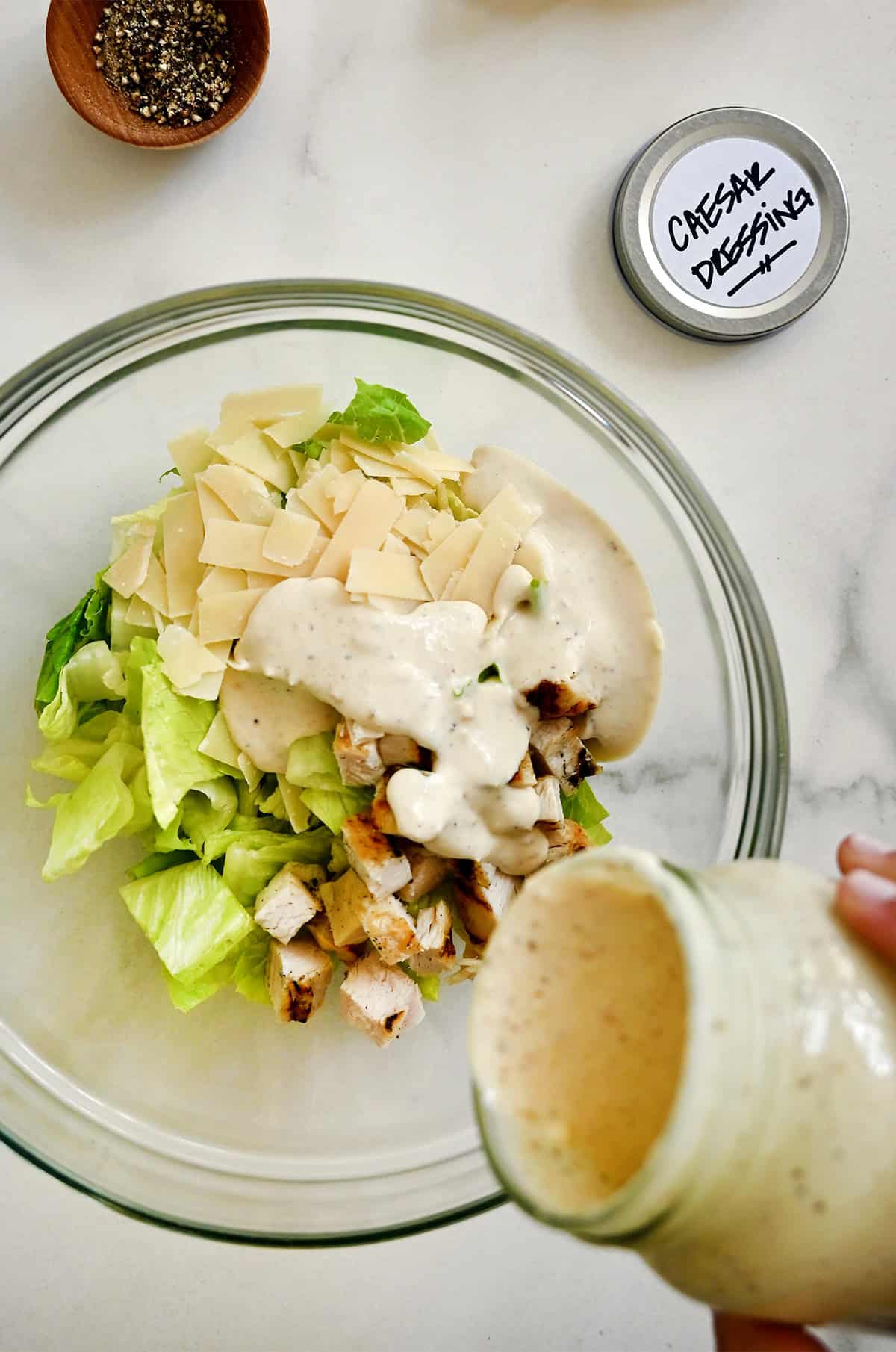 Adding homemade Caesar salad dressing to a large mixing bowl with chopped chicken, romaine lettuce and shaved Parmesan cheese.