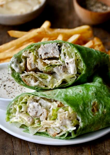 Chicken Caesar salad in a spinach wrap on a plate with French fries and a small bowl containing Caesar dressing.