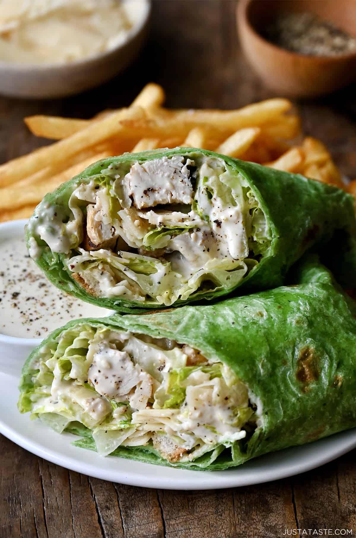 Chicken Caesar salad in a spinach wrap on a plate with French fries and a small bowl containing Caesar dressing.