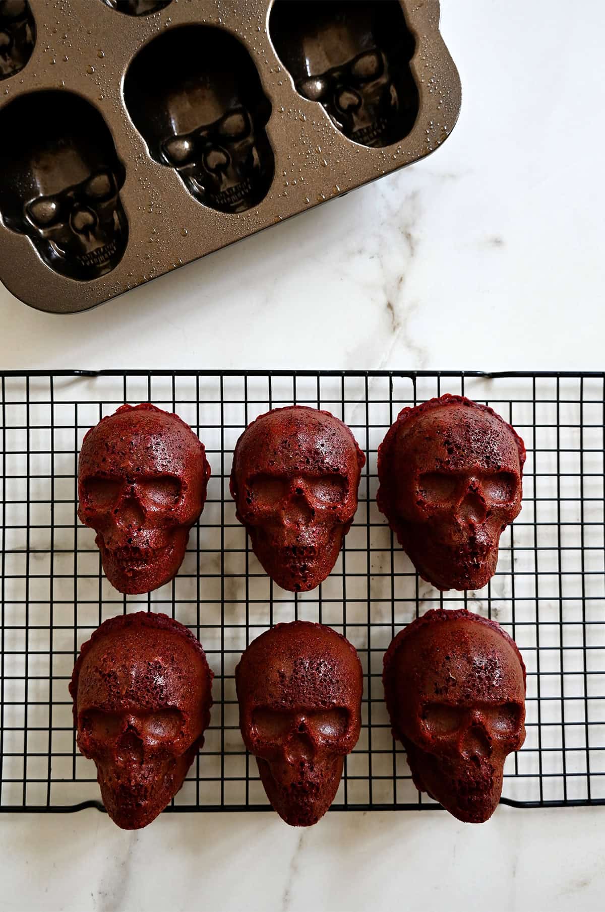 Mini red velvet skull cakes cooling on a wire rack next to a skull cake pan.