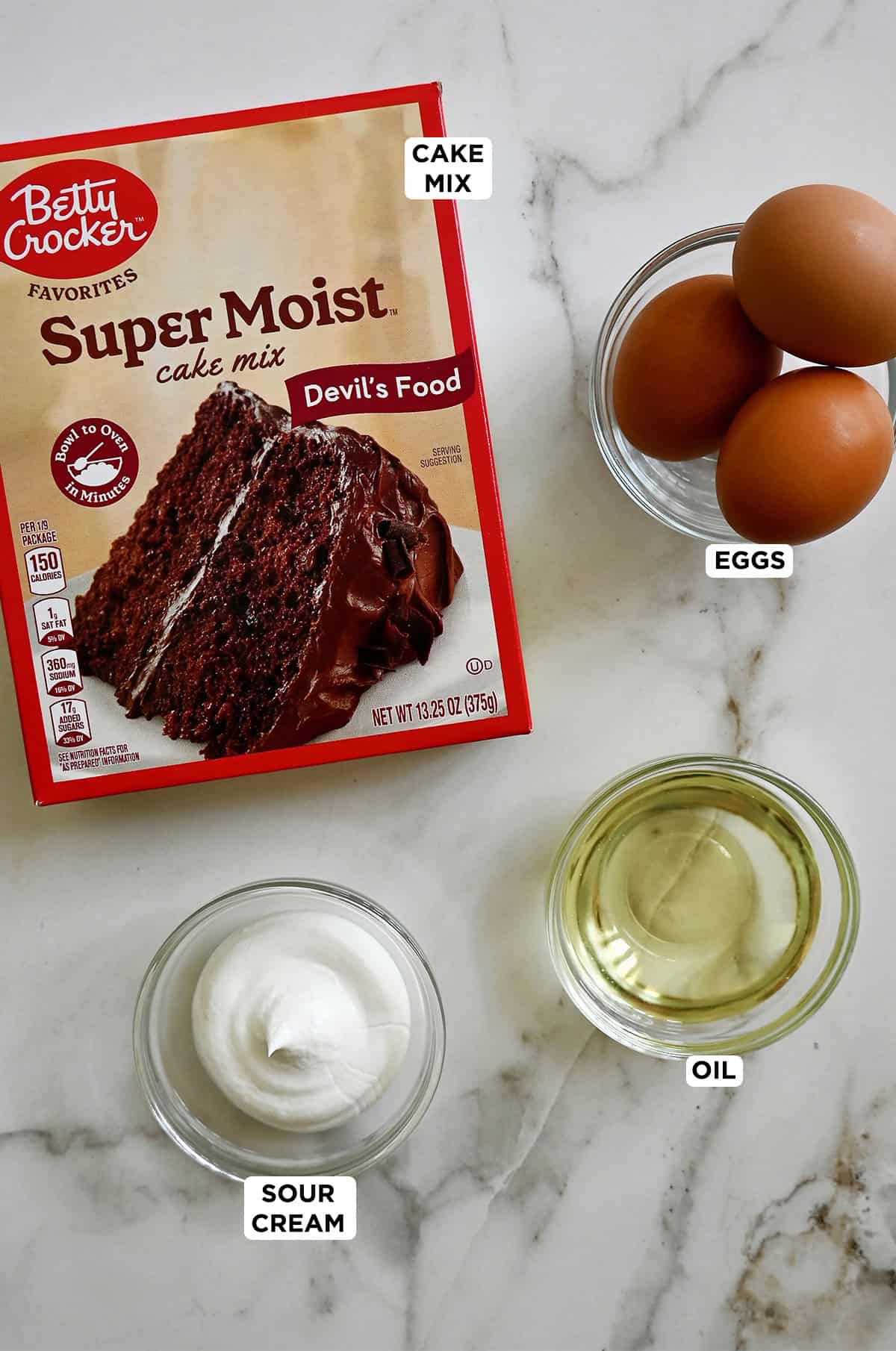 A box of devil's food cake mix next to three bowls containing eggs, vegetable oil and sour cream.