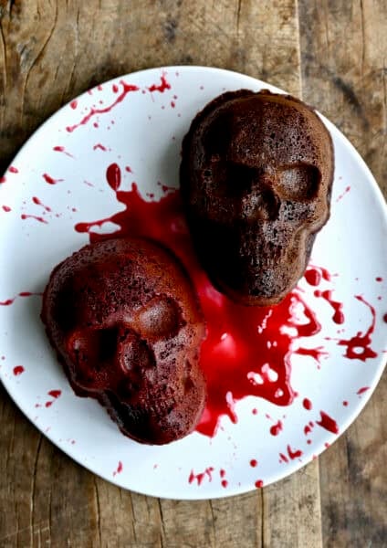 Two mini Halloween skull cakes on a plate, splattered with raspberry sauce.